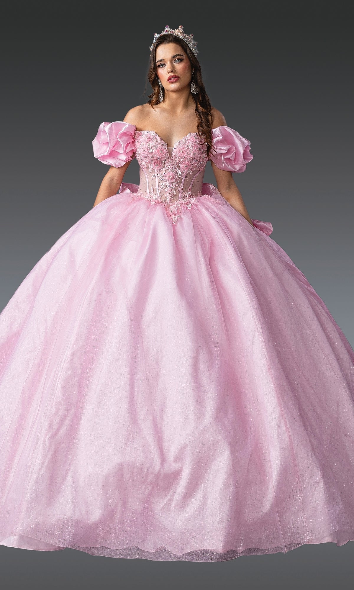 A woman in the Quinceanera Dress 1994 by Dancing Queen, a light pink off-the-shoulder gown with floral details and a sparkling tiara, stands against a neutral background.
